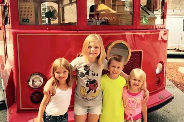 Four children smiling in front of a red vintage trolley.
