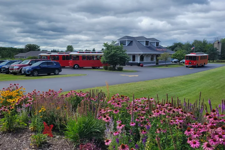 Red trolleys parked by a building, with colorful flowers in the foreground and cloudy sky.